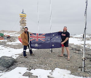 Two men stand holding Australian flag, man on right is in thongs, shorts and t-shirt. They are outside on snow covered rocky ground beside flag poles and the casey sign