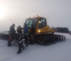 Wilkins team of four standing around groomer front blade having a discussion, in fog and whiteout conditions