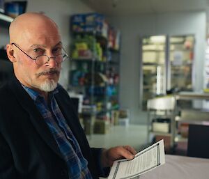 Close up of man in jacket, wearing glasses on nose, with eyebrows drawn on, holding passenger list. In the background the kitchen