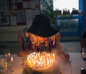 Man dressed as woman with large floppy black hat sits as table and blows out candles on a birthday cake