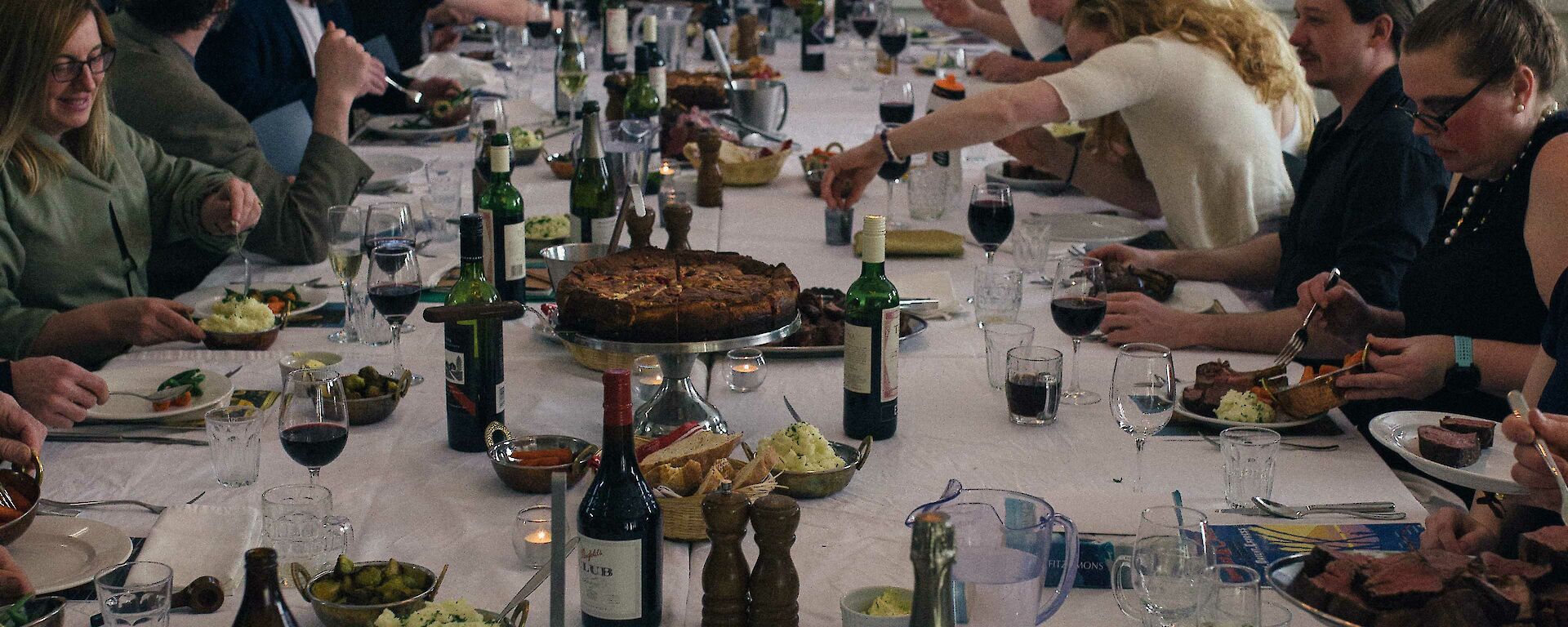 Long table set for dinner, laden with food and drinks, with old fashioned candelabra above