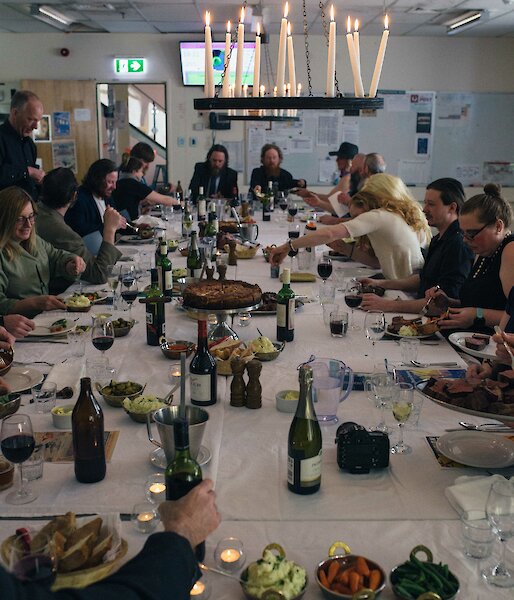 Long table set for dinner, laden with food and drinks, with old fashioned candelabra above