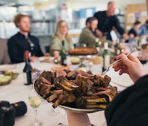 Close up of tray of lamb racks with people at table in background