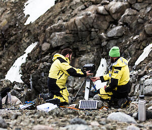 Two men kneeling and adjusting camera box sitting on tripod, surrounded by steep rocky landscape