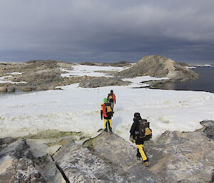 Men walking in single file, carrying field packs, walking across snow covered ground towards rocky hill in distance. Distant penguin rookery in middle of picture