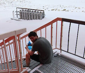Man sits on outside metal stairs looking sweaty and tired, in background snow covered ground to horizen