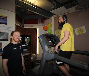 On left man standing beside treadmill smiles at camera, on right man walking on treadmill, wearing headphones, smiles at other man. In background are posters on wall, balloons, television with TV show running and Camp Quality pictures