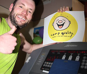 Man stands besides treadmill’s display which shows 42.2km travelled, and holds up Camp Quality sign