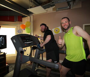 Man on treadmill takes big exaggerated step, second man beside treadmill smiles at camera and gives the thumbs up