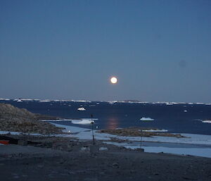 Landscape of rocky ground, station services in foreground, then sea with small ice bergs, and middle of picture just above the horizen is the full moon