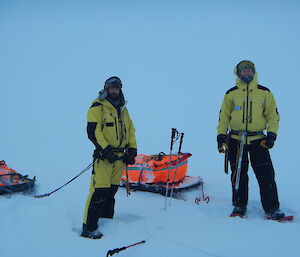 Two men dressed in winter gear with sleds attached to harnesses on waists, on sea-ice