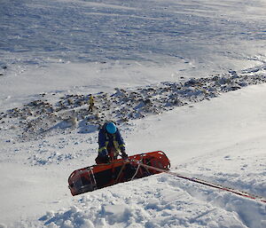 Stretcher holding person being lowered over edge of ice cliff, attached by rope, with person on side of stretcher assisting it move down the slope