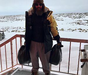 Man standing on outside veranda holding a bag of rubbish in each hand