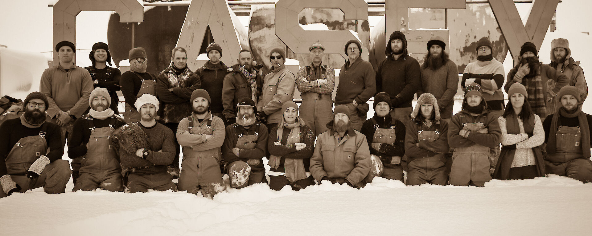 Sepia photo of group of 26 expeditioners, back row standing and front row kneeling, in front of large metal CASEY sign on low fuel farm tanks