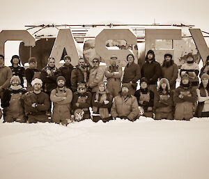 Sepia photo of group of 26 expeditioners, back row standing and front row kneeling, in front of large metal CASEY sign on low fuel farm tanks