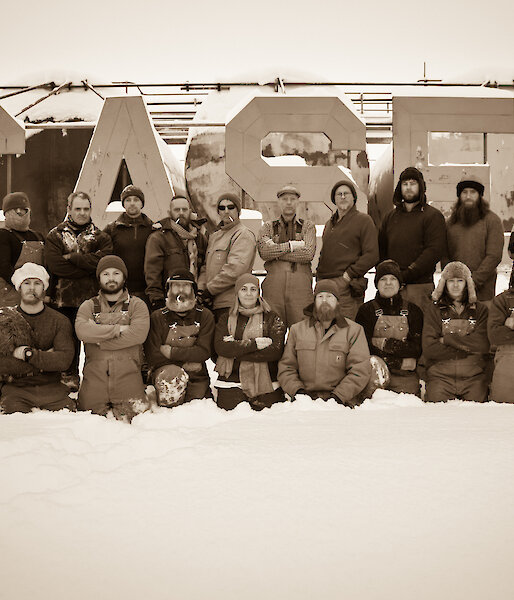 Sepia photo of group of 26 expeditioners, back row standing and front row kneeling, in front of large metal CASEY sign on low fuel farm tanks