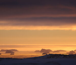 Orange glow of sunset is picking up hazy icebergs in the distance on the horizen, foreground snow covered outcrop, above grey clouds