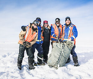 Sunny blue sky day, group of five people in winter gear gathered together smiling at camera behind large parachute bag
