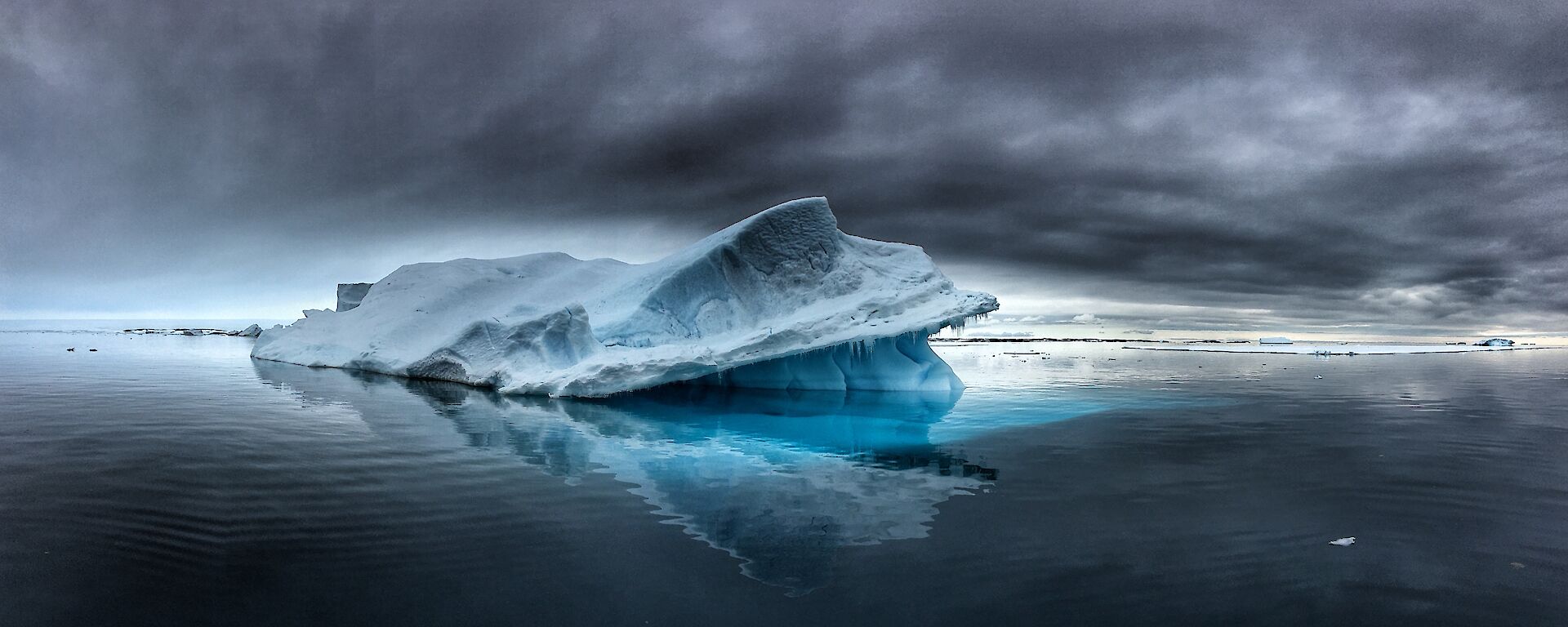 Panoramic view of iceberg slighly toppled over with calm grey sea matching ominous grey clouds above