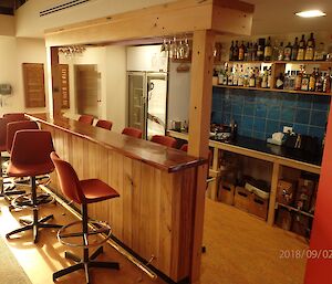 Close up of bar area, showing fridge, benches and shelving for bottles and bar divide with bar stools