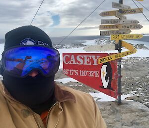 Man standing in front of the Casey signpost in his googles, beanie, face cover and heavy jacket with the rocks and bay in background