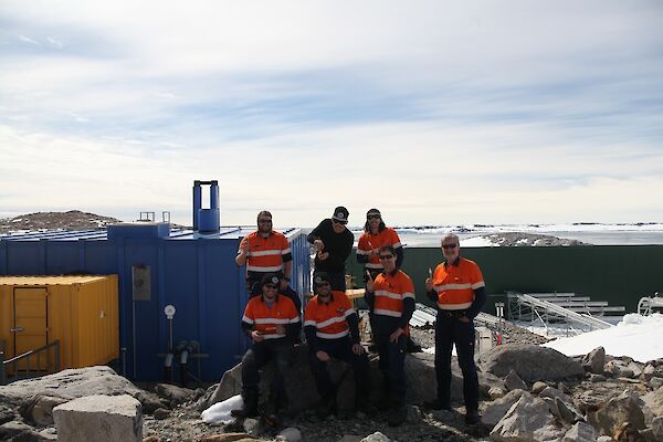 The electrical team standing near the Casey Emergency Power House (EPH, Blue building) with their “magic” test sticks and Newcomb Bay in the background.