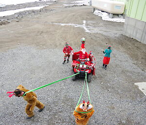 Grown men in elves costumes pull another grown man in a santa costume in a sled