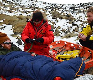 Two expeditioners provide first aid to a third expeditioner lying on the ground in a search and rescue exercise