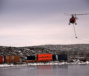 A helicopter sling carrying a rope beneath its skids approaches a container wharf at Davis