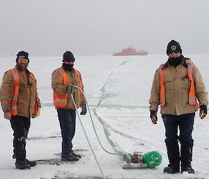 Three expeditioners stand in foreground with ship and fuel hose on sea ice in background