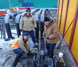 Three expeditioners facing camera next to pipe work
