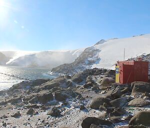 A red field hut positioned above a frozen water source and steep cliffs giving way to a plateau