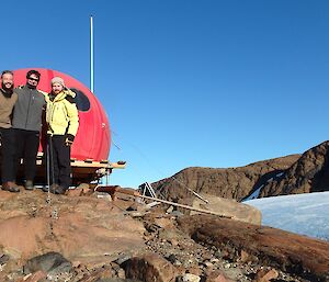 Three expeditioners stand in front of a field hut.