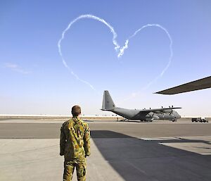 Man in camouflage standing in foreground. In background a C130 hercules aircraft with a cloud shaped heart above it