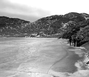 Three expeditioners walk on the shore next to a frozen body of water