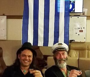 Two expeditioners sip a drink beneath a Greek flag