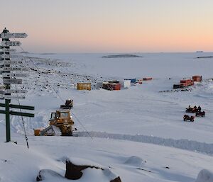 Snow groomers descend down the road from Davis whilst quad bike riders wait for the road to be cleared in background. In foreground Davis station signpost