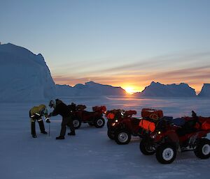 Expeditioners drilling sea ice with sun setting in background