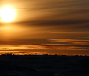 Impressive sunset with large sun in top left of shot, and a streaky, fiery sky above indiscernible water and icebergs, merely silhouettes in the distance