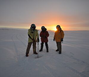 Expeditioners stand in front of sunrise conducting a sea ice drill