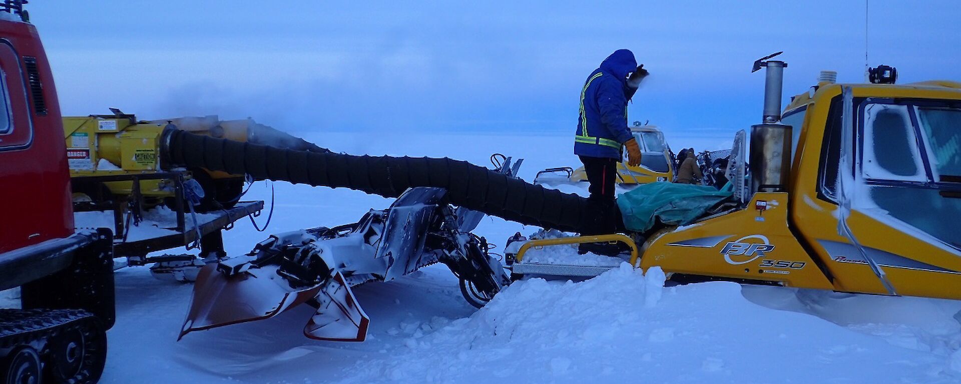 Expeditioner standing above a snow groomer as a heater with an extension tube defrosts the engine bay