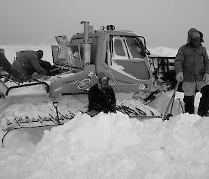Three expeditioners in place beside and on a snow groomer whilst excavating it from snow drift