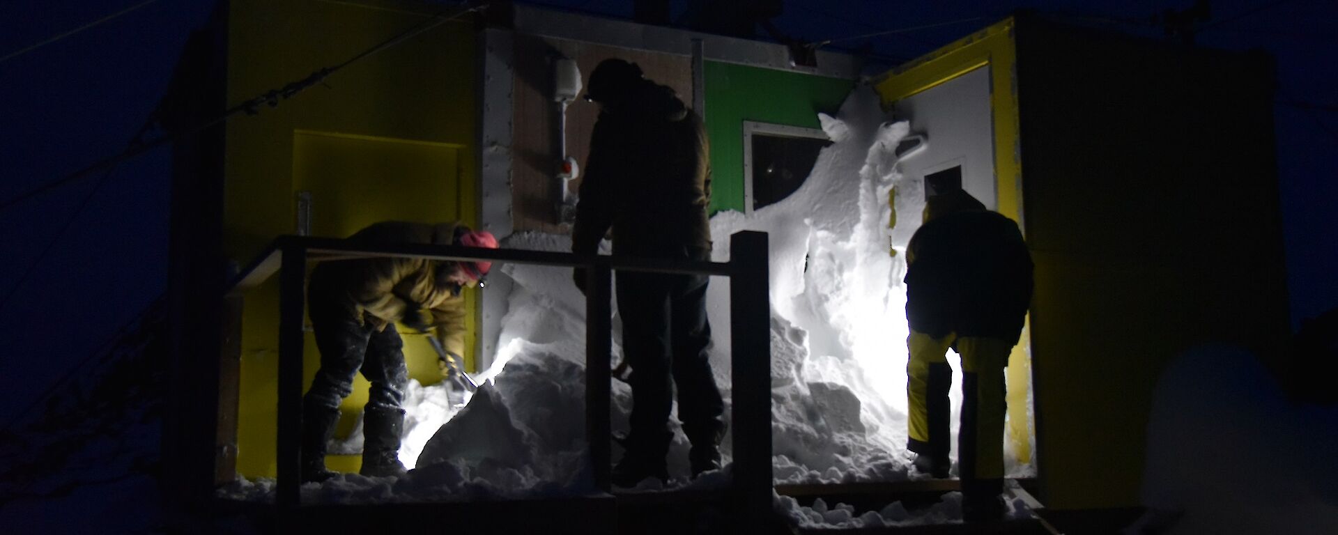 Three expeditioners illuminated by torches dig snow out of the entrance of Platcha Hut