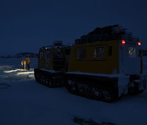 Yellow tracked vehicle in foreground illuminates two expeditioners drilling sea ice in its headlights