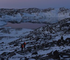 Three expeditioners wearing backpacks walk through snow and rocks towards a lake surrounded by snow covered hills in fading light