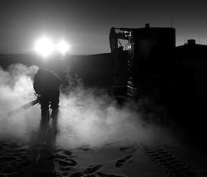 Expeditioner silhouetted in a vehicles lights applying a chainsaw to the sea ice.