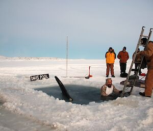 Expeditioner dressed in a leopard seal costume descends a ladder into a square hole containing cold sea water.