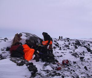 Several expeditioners sheltering behind a large boulder as other expeditioners arrive on scene in background