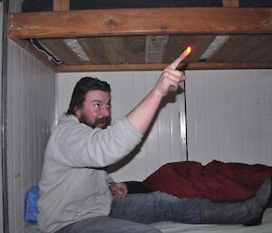 Expeditioner sitting on bunk bed in field hut with finger illuminated by torch.
