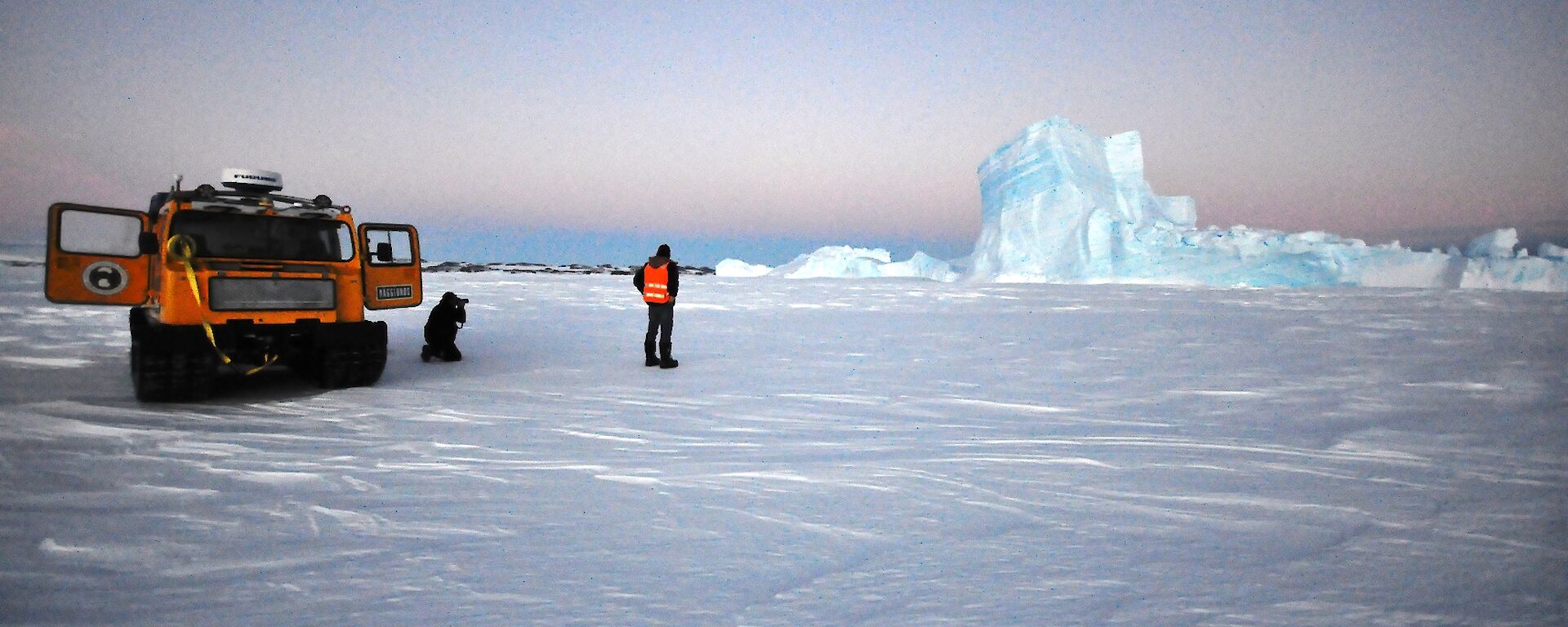 Two expeditioners beside a tracked vehicle looking right towards an iceberg encased in sea ice